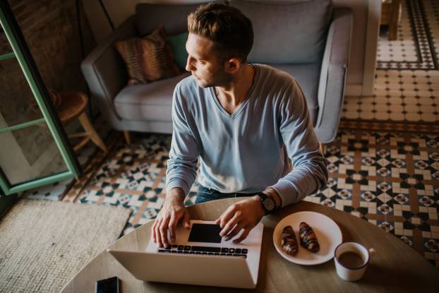 Handsome coworker man working at living room at home. Man sitting at wooden table using laptop and mobile phone. Blurred background.