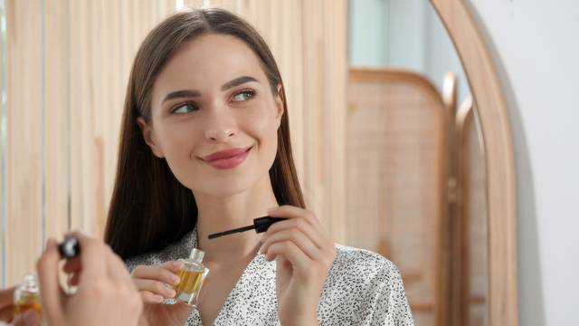 Young woman with eyelash oil near mirror indoors, space for text