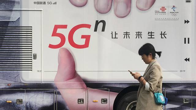 Woman using her mobile phone walks past a vehicle covered in a China Unicom 5G advertisement in Beijing