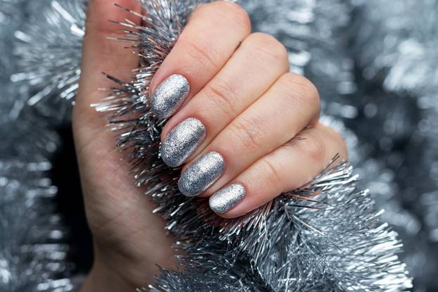 Female hand with beautiful holiday manicure - silver glittered nails on Christmas tinsel background. Selective focus. Closeup view. Blurred background