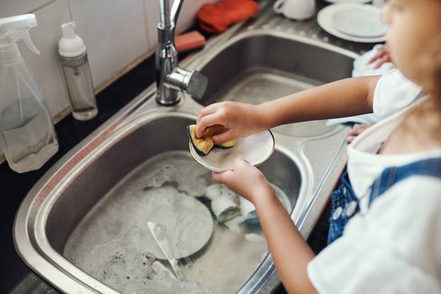 Girl, water and learning to wash dishes in kitchen, sanitary and prevent bacteria or germs. Female person, child and soap for cleaning mess in home, housework and kid helping for responsibility