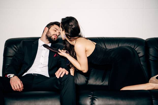 attractive girl in black dress holding tie of passionate man in suit sitting on sofa 