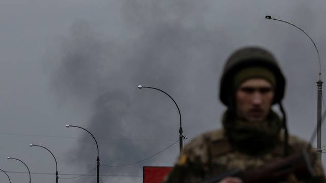 Smoke rises as a service member of the Ukrainian armed forces stands by the only escape route used by locals to evacuate from the town of Irpin, after days of heavy shelling, while Russian troops advance towards the capital, in Irpin, near Kyiv