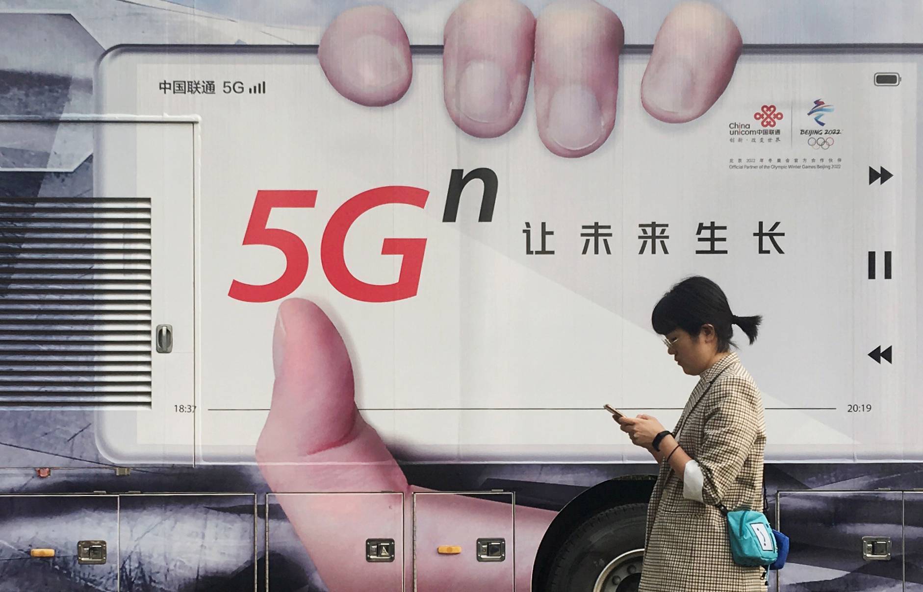 Woman using her mobile phone walks past a vehicle covered in a China Unicom 5G advertisement in Beijing