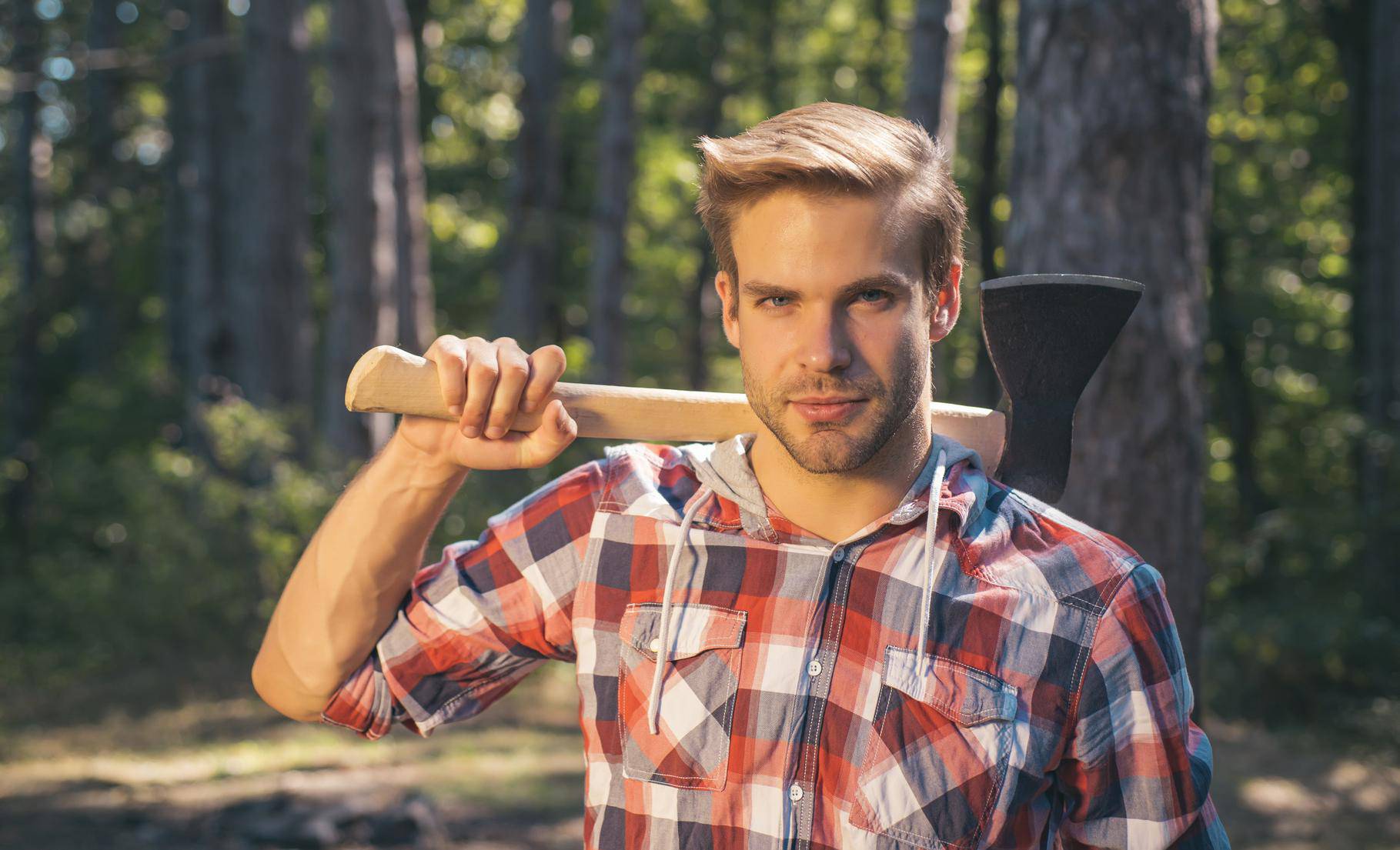 Lumberjack carries axe on shoulder. Deforestation. Handsome man with axe. Lumberjack in the woods with an ax.