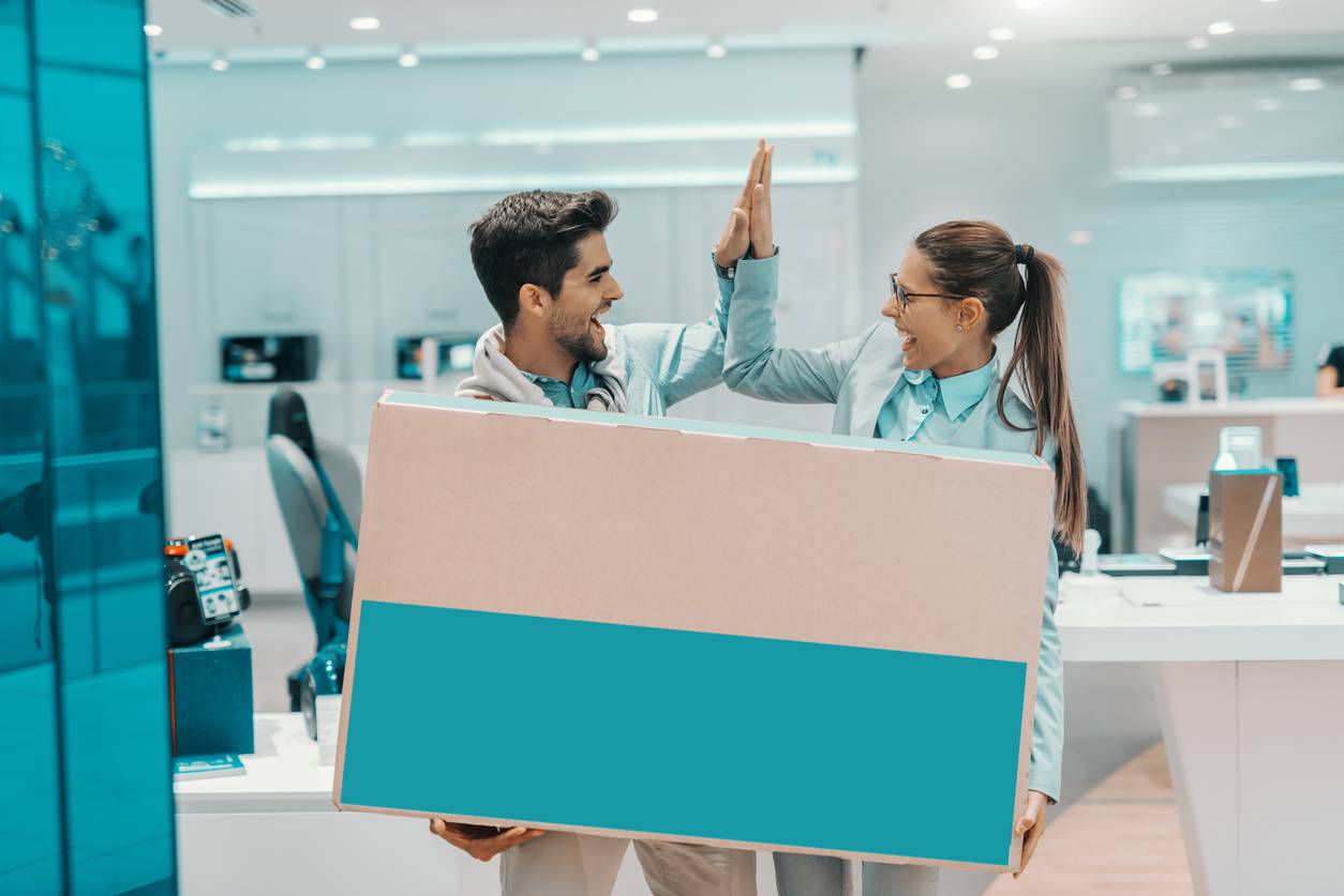 Smiling happy multicultural couple dressed elegant holding box with new plasma tv and giving high five to each other while standing in tech store.