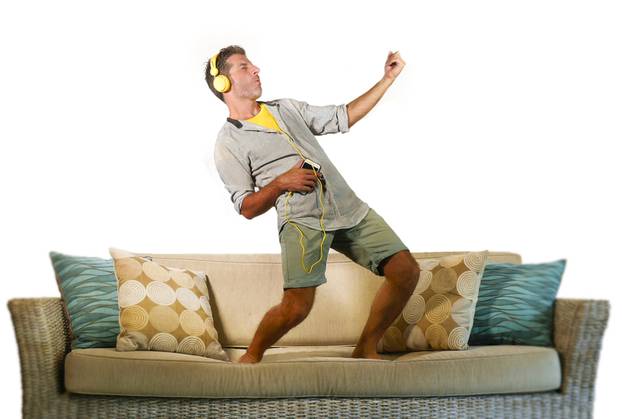 young happy and excited man jumping on sofa couch listening to music with mobile phone and headphones playing air guitar crazy having fun at home