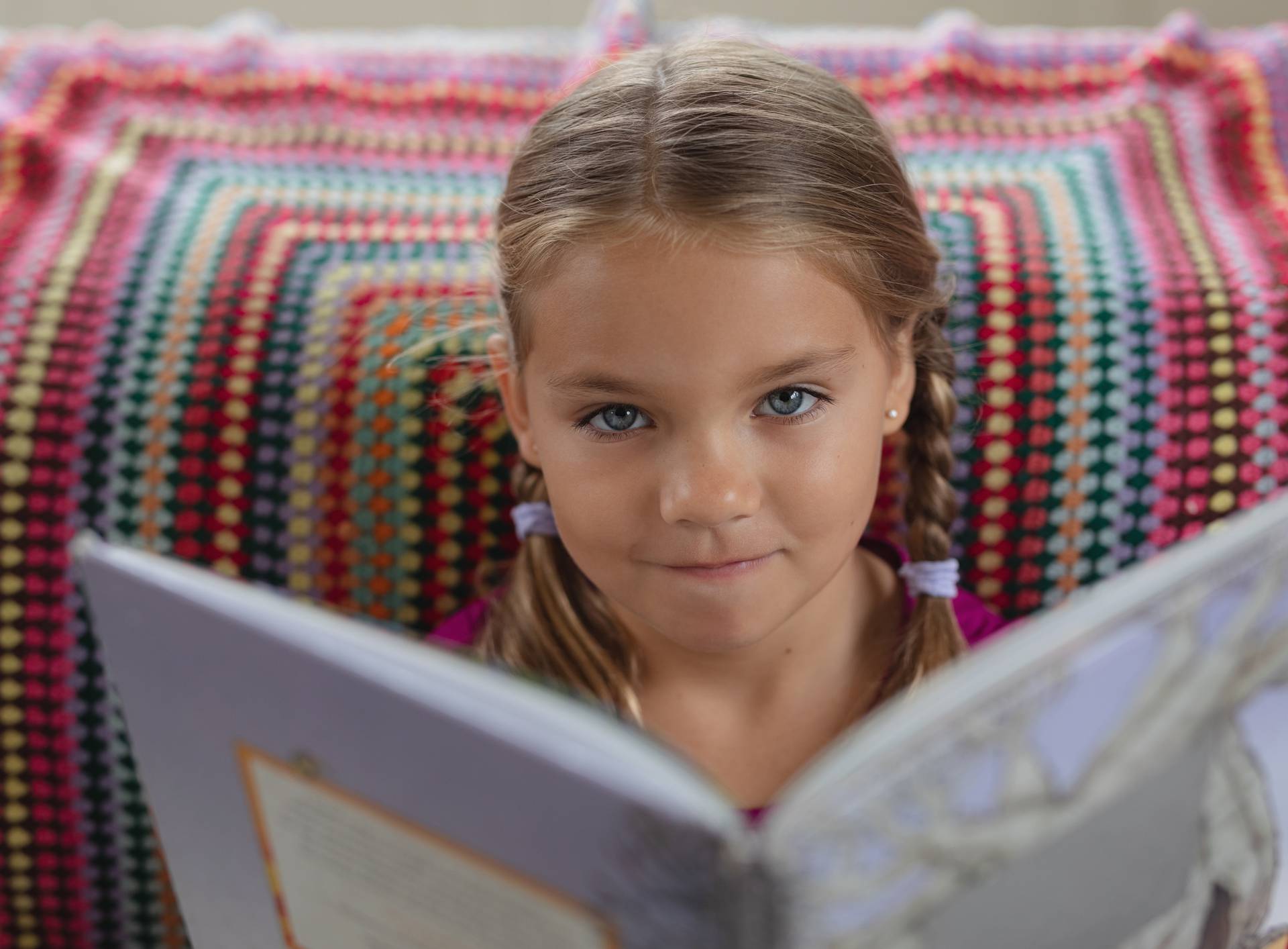 Font view of cute Caucasian girl looking at camera while reading a book on sofa in a comfortable home