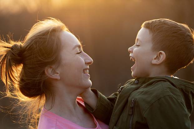 Close up of a lovely mother and her son having fun outdoor. Little cute kid holded by her mom in the arms which is laughing against sunset. Mother's day concept