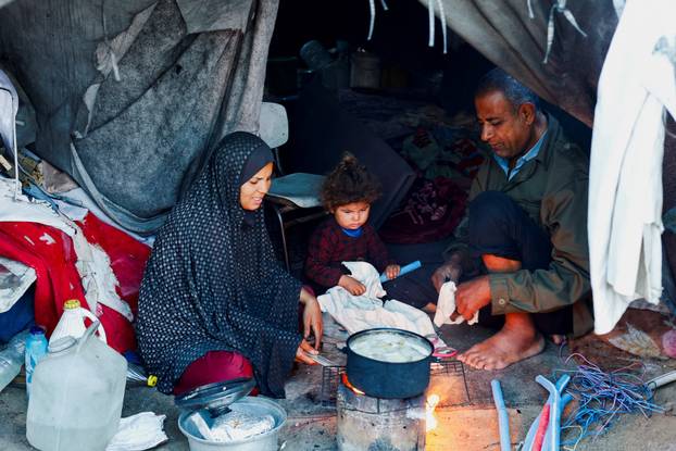 A Palestinian family prepares food over a fire stove in front of their tent in a camp on a rainy day in Nuseirat