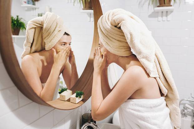 Young happy woman in towel applying organic face scrub and looking at round mirror in stylish bathroom. Girl making facial massage, peeling and cleaning skin on face. Skin Care and Hygiene