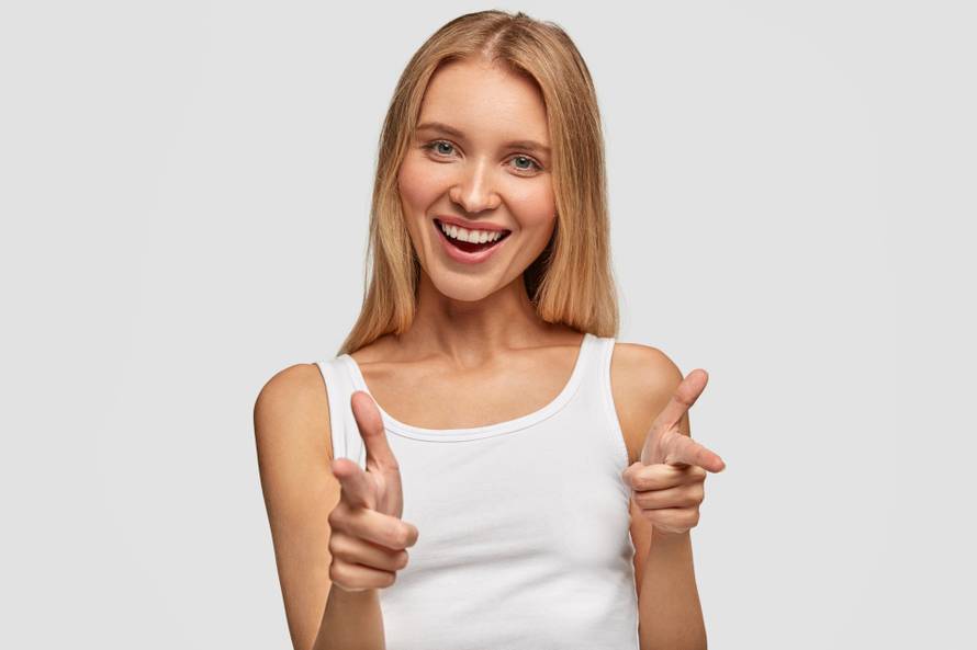 Portrait of happy cute blonde female with joyful expression, points at you with fore fingers directly at camera, chooses companions for entertainment, dressed casually, isolated over white background