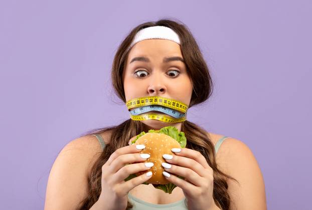 Shocked millennial european woman plus size in sports uniform with closed mouth with measuring tape look at burger