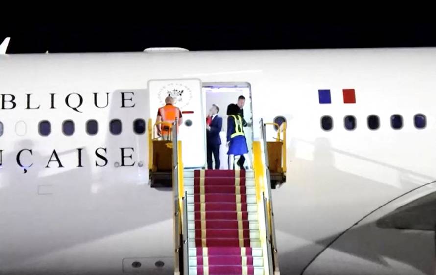 French President Emmanuel Macron stands near the door of a plane, as a hand appears and pushes his face away, in Hanoi