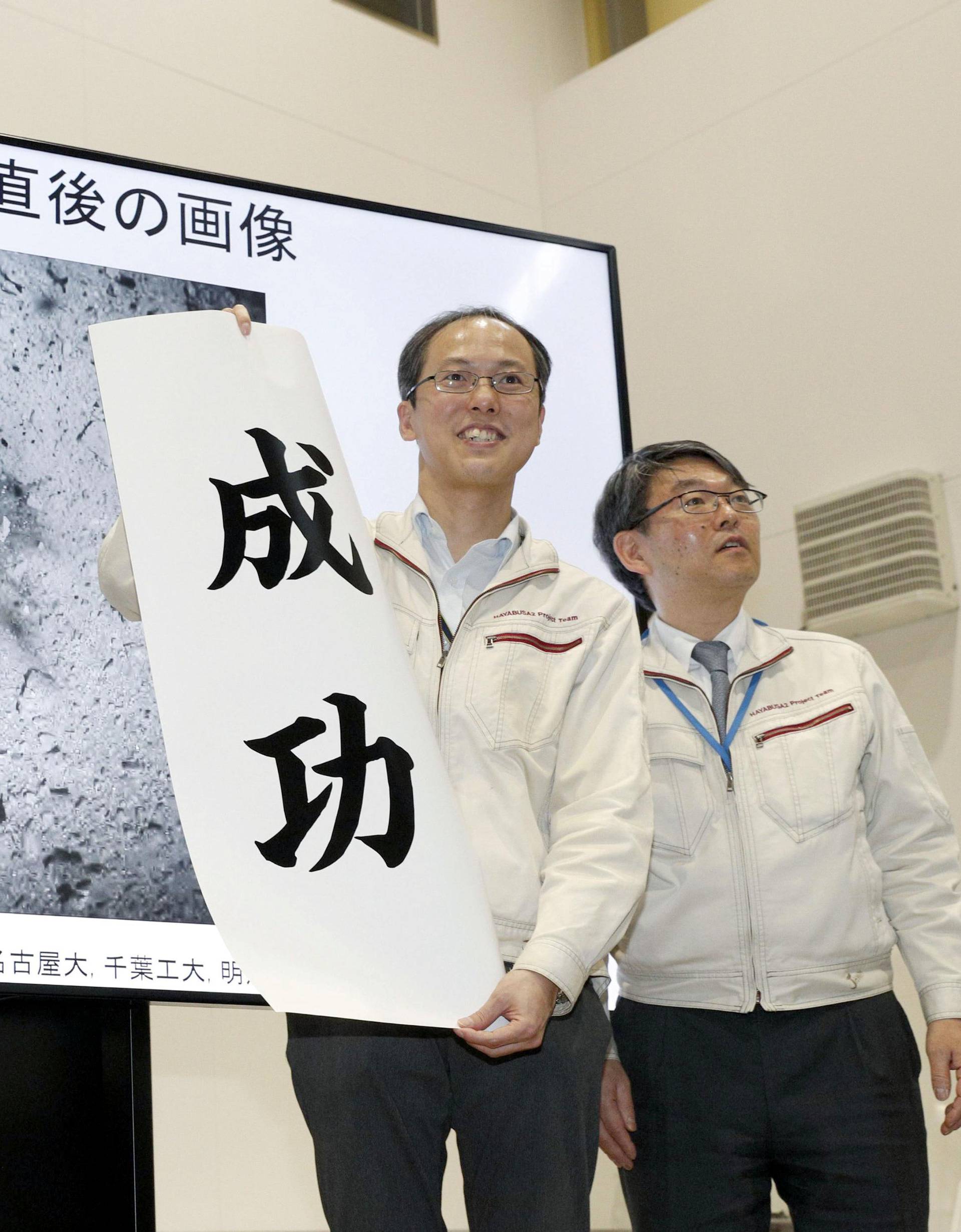JAXA associate professor Yuichi Tsuda holds a banner reading 'success' in front of an image of the Hayabusa 2 space probe's landing on the Ryugu asteroid in Sagamihara