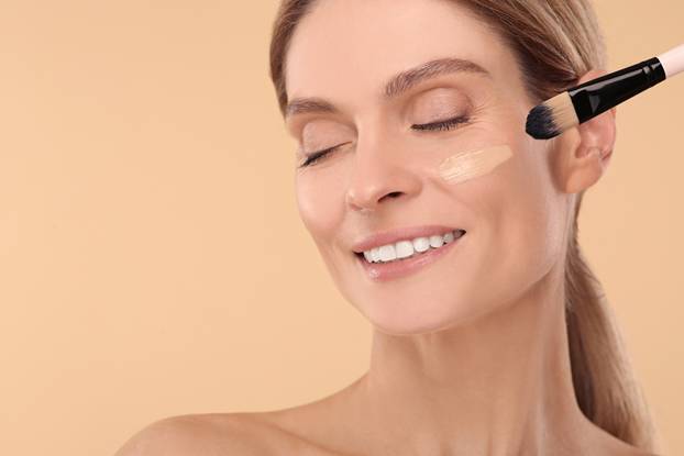 Woman applying foundation on face with brush against beige backg