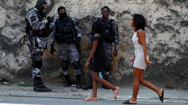 Residents walk near police officers during an operation against drug gangs at Fallet slum in Rio de Janeiro