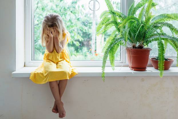 5 years old blonde girl sit on the windowsill and cry covering her face with her hands