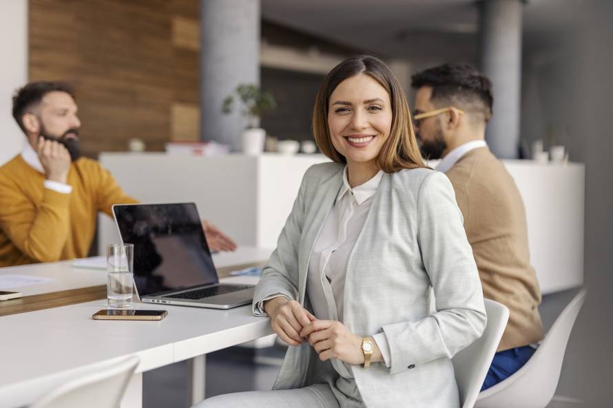 Portrait of happy businesswoman in formal wear sitting in boardroom with colleagues during the meeting and smiling at camera.