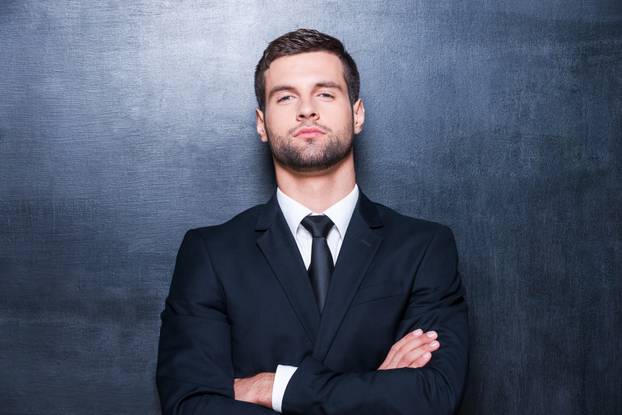 Confident business. Handsome young man in shirt and tie looking at camera and keeping arms crossed while standing against blackboard