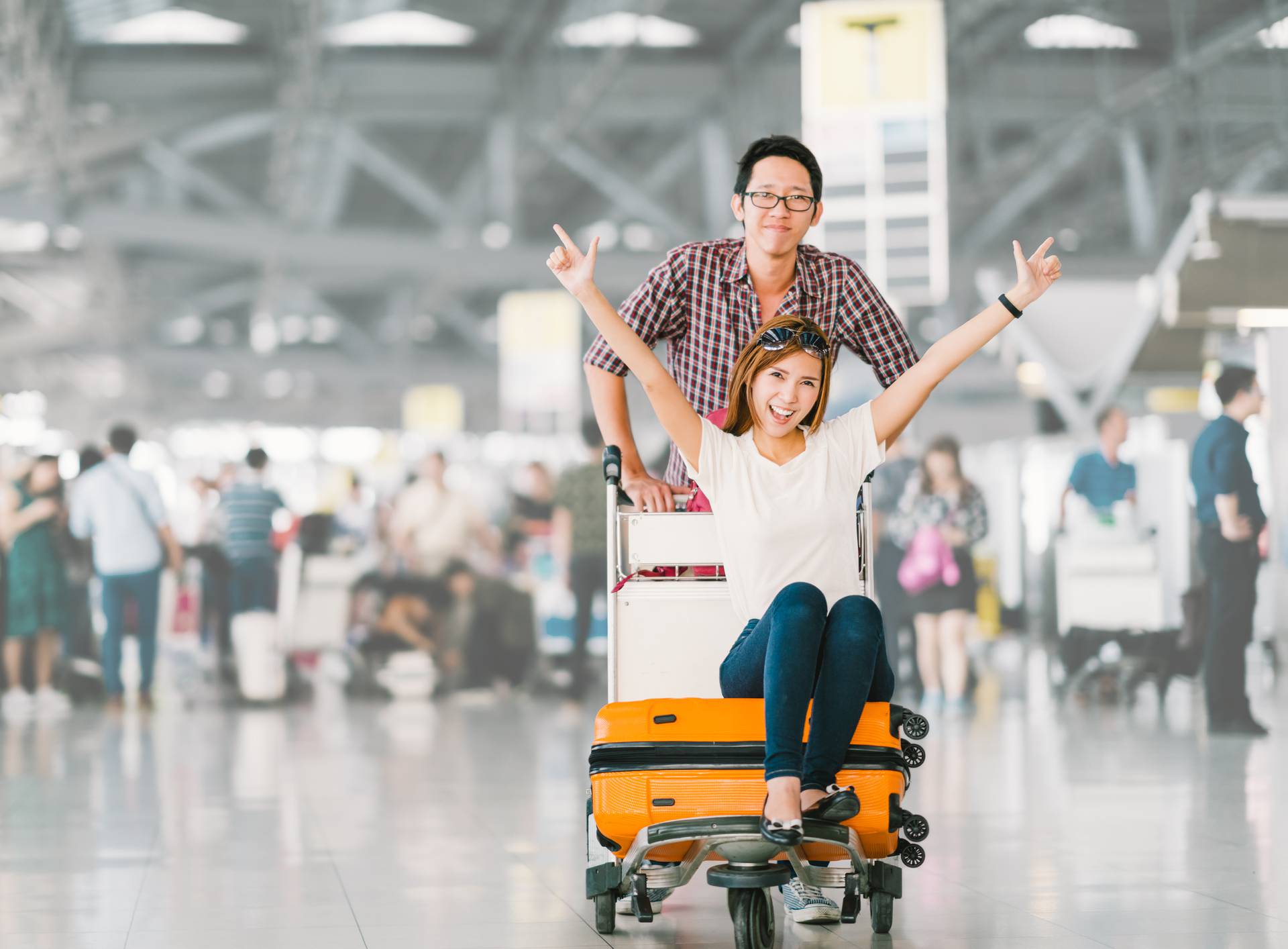 Young Asian tourist couple happy and excited together for the trip, girlfriend sitting and cheering on baggage trolley or luggage cart. Holiday vacation travelling abroad concept, with copy space