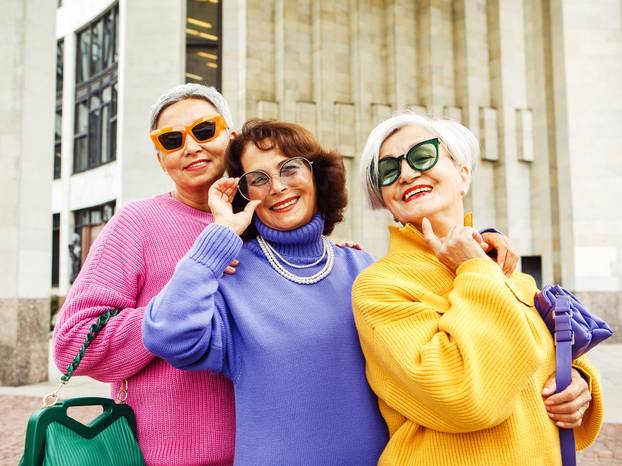 Three happy cheerful pensioner girlfriends in bright sweaters and sunglasses walk together