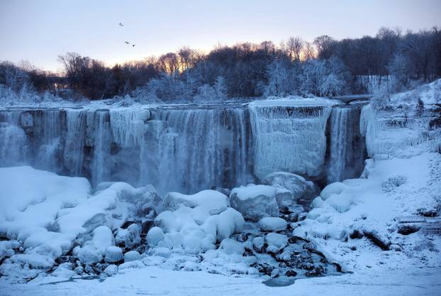 A group of birds fly past ice and water flowing over the American Falls, viewed from the Canadian side in Niagara Falls,