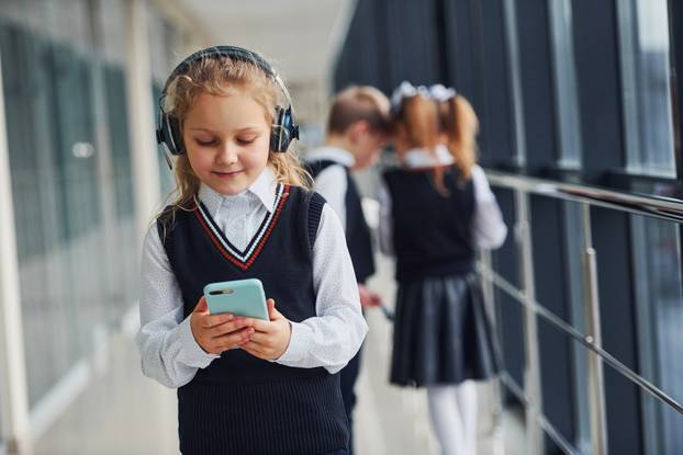 Little girl with phone and headphones standing if front of school kids in uniform that together in corridor. Conception of education