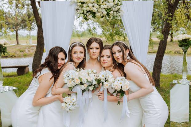 Group portrait of the bride and bridesmaids. Bride in a wedding dress and bridesmaids in white dresses and holding stylish bouquets on the wedding day against the background of the lake.