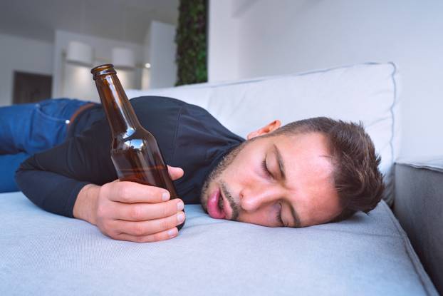 Drunk young man with open mouth and bottle of beer sleeping on the sofa at home.