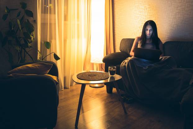 Young woman uses laptop watch course, checks email news online sitting on sofa. Beautiful girl working on a computer, watching a webinar at home in cozy blanket in winter by lamp