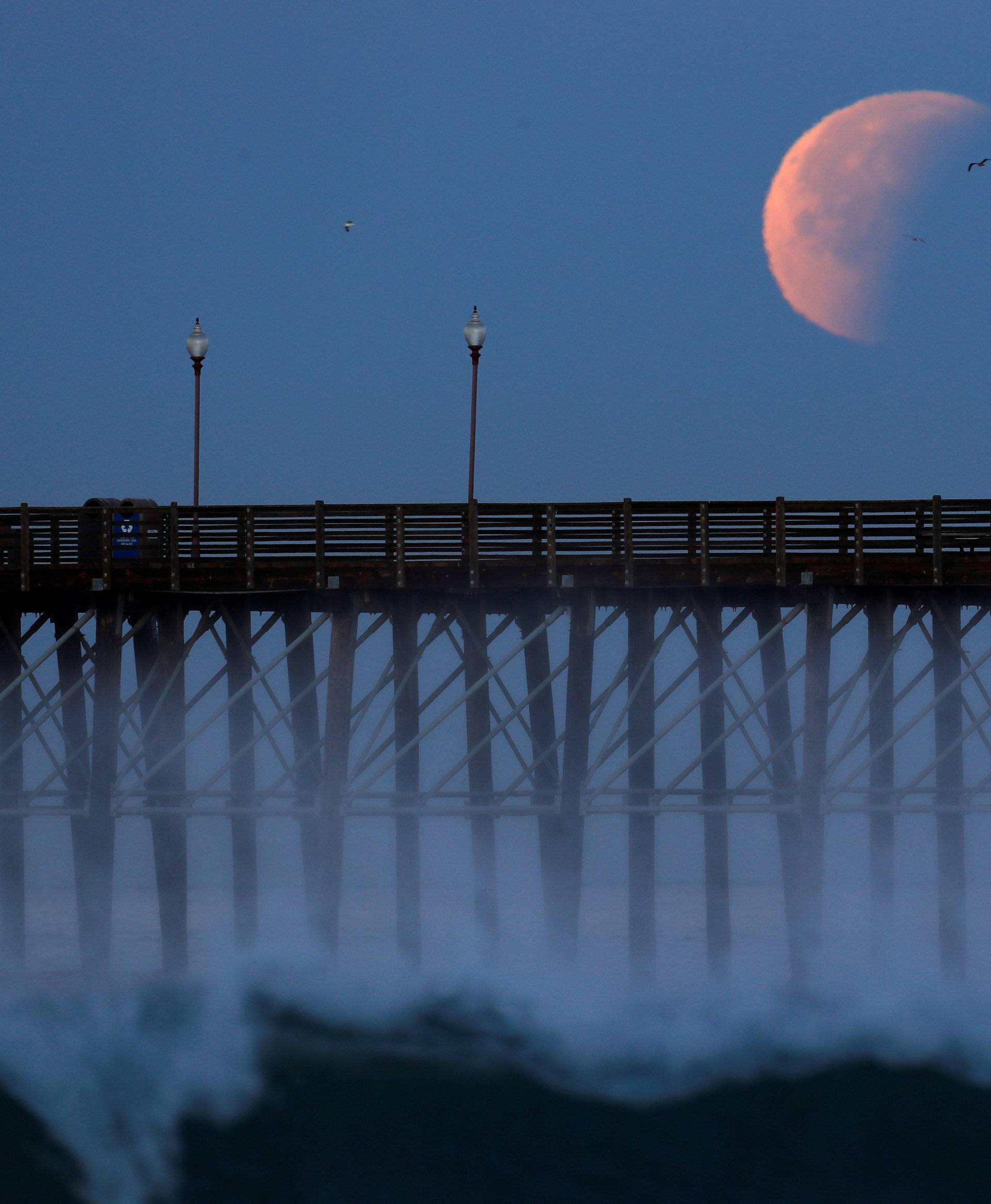 A Blue Moon comes out of a lunar eclipse as it sets past an ocean pier in Oceanside, California