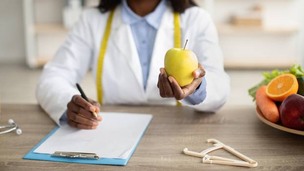 Black female doctor making prescription or writing diet plan and holding apple, sitting at desk with fruits and caliper