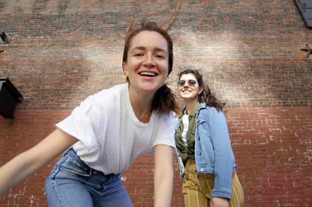 Portrait of two young millennial women, wearing casual clothing, move playfully in front of a brick wall.