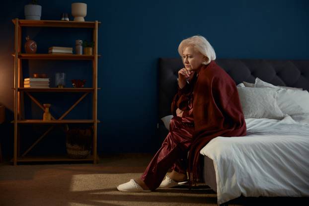Elderly woman wearing pajamas sitting on bed feeling loneliness