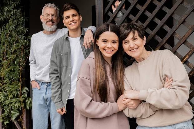 Smiling teenage girl holding hand of middle aged mother while looking at camera near blurred father and brother during parents day celebration on porch of house, parent-child relationship concept