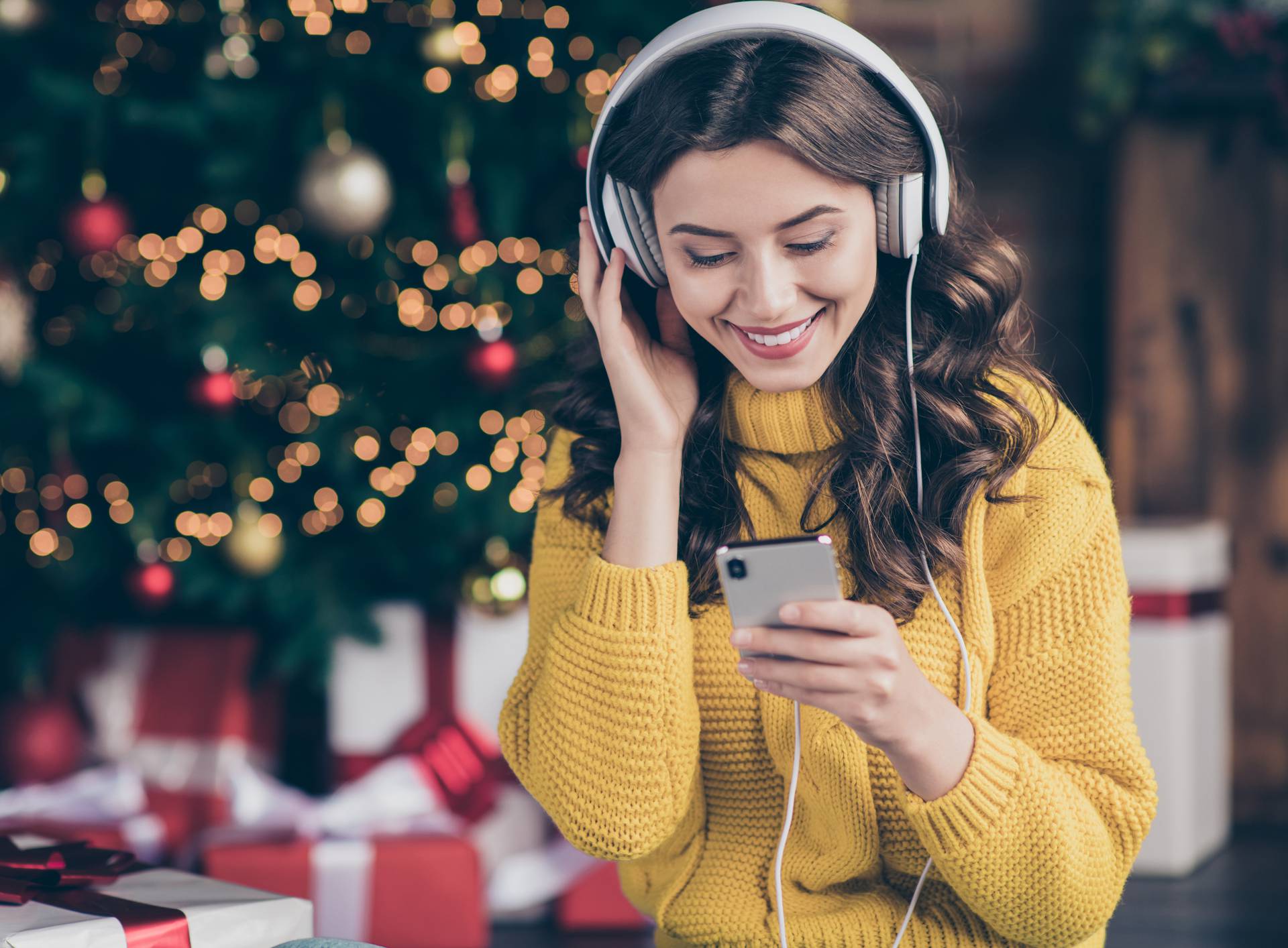 Photo of charming beautiful wavy curly haired girl listening to music wearing yellow pullover staring at telephone screen