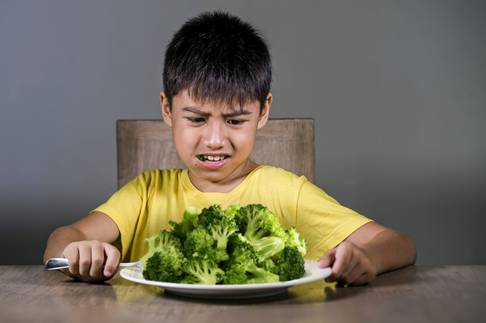 7 or 8 years old upset and disgusted Asian kid sitting on table in front of broccoli plate looking unhappy rejecting the fresh food in child hate green vegetables concept