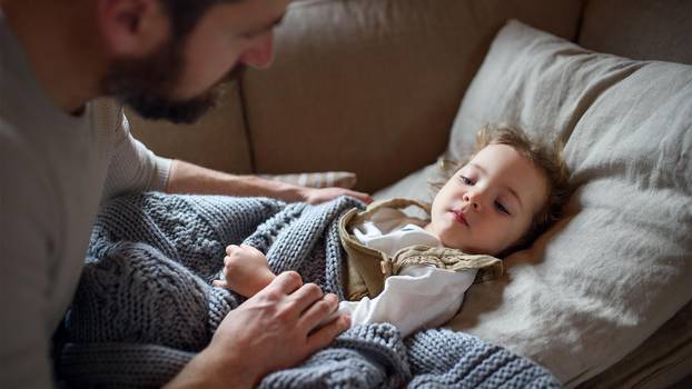 Father checking on small sick daughter lying in bed indoors at home.