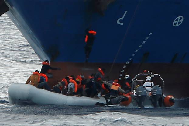 Migrants in a rubber dinghy fall into the sea alongside a RHIB of the Malta-based NGO Migrant Offshore Aid Station, as a migrant climbs to the anchor of the Panama-registered ship Tuna 1 and one hangs onto its bow