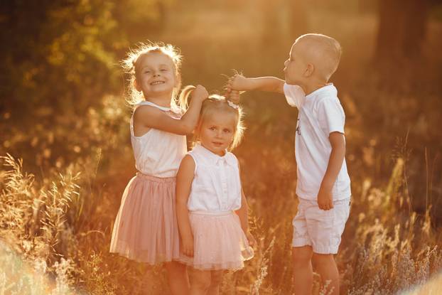 two sisters and brother are playing, hugging in the field on summer. children playing outdoors and smiling on sunset. happy family. happy childhood