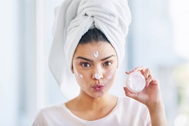 Gotta fight those signs of aging. Portrait of a beautiful young woman applying moisturizer to her skin in the bathroom at home.