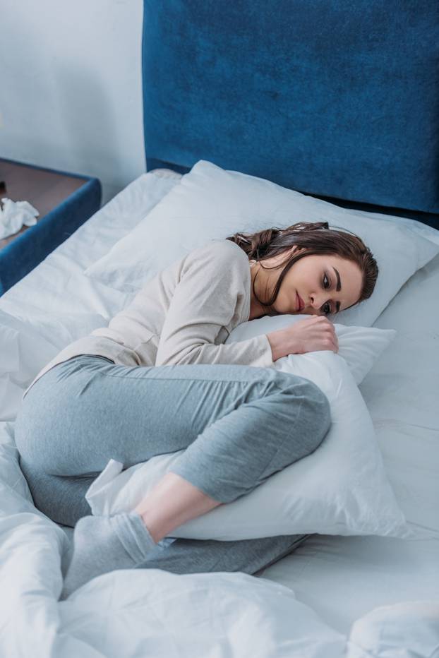 sad woman in pajamas holding pillow and lying in bed at home