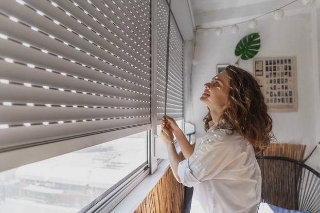 Young woman opening roller blind shutters on the balcony on a summer day