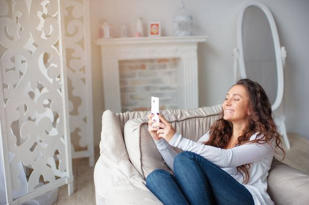 Young attractive woman taking picture with her white smartphone sitting on sofa at home.
