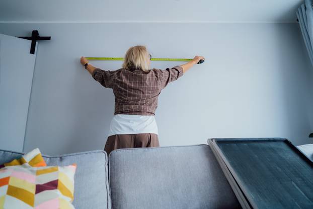 Back view Middle aged woman doing measuring with a measure tape on the wall. Lady wants to put a picture on the wall at home. Housekeeping work. Doing repair herself. DIY, equality in work concept.