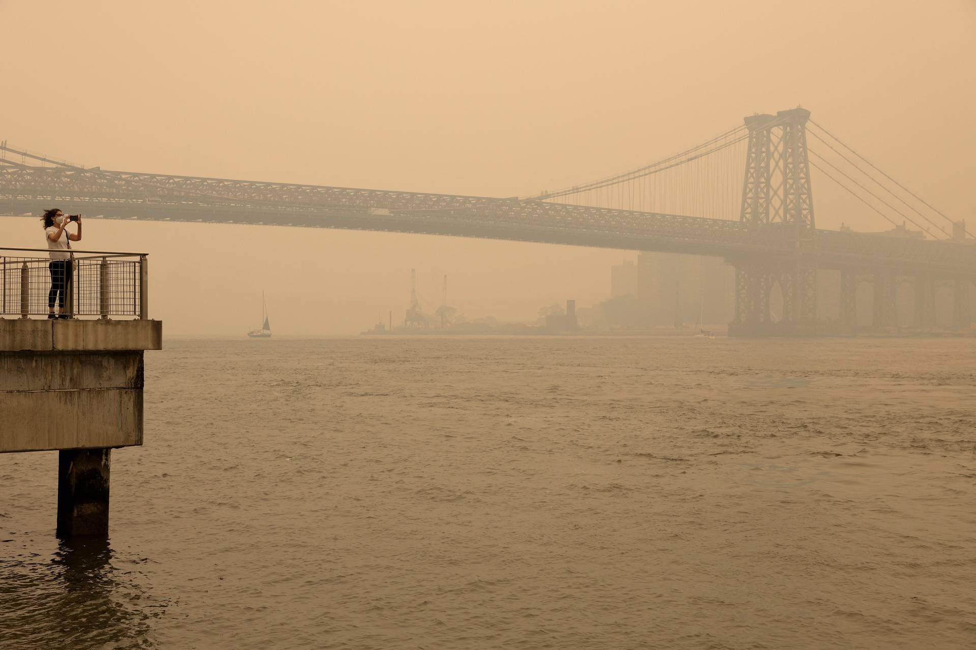 A person wearing a face mask takes a picture of the Manhattan skyline while it’s covered in haze and smoke caused by wildfires in Canada, in Brooklyn, New York