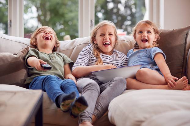 Three,Children,Sitting,On,Sofa,At,Home,Laughing,And,Watching