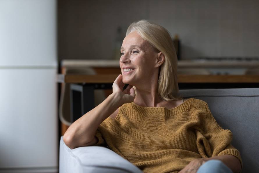 Attractive middle-age woman daydreaming seated on sofa in studio apartment