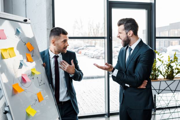 angry businessmen screaming at coworker near white board in modern office 